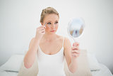 Natural serious blonde holding mirror and using tweezers
