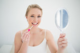 Natural smiling blonde holding mirror and applying lip gloss