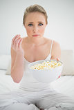 Natural frowning blonde holding bowl of popcorn on bed