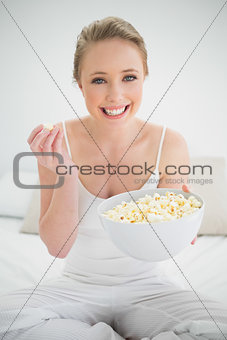Natural cheerful blonde holding bowl of popcorn on bed