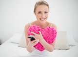 Natural cheerful blonde holding heart pillow and smartphone