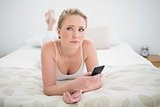 Natural pensive blonde lying on bed and using smartphone