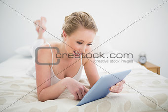 Natural smiling blonde lying on bed using tablet