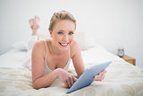 Natural smiling blonde lying on bed holding tablet