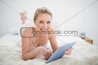 Natural smiling blonde lying on bed holding tablet