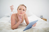 Natural thoughtful blonde lying on bed holding tablet