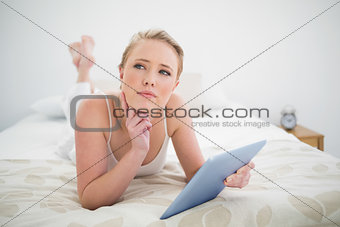 Natural thoughtful blonde lying on bed holding tablet