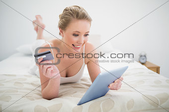 Natural happy blonde lying on bed holding tablet and credit card