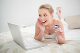 Natural excited blonde holding credit card and looking at laptop