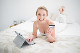 Smiling natural blonde lying on bed and using tablet and credit card