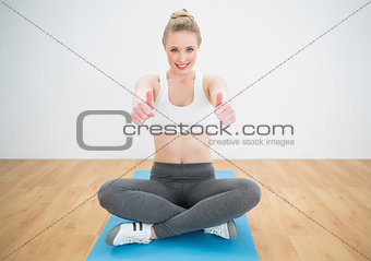 Smiling sporty blonde sitting cross legged on exercise mat showing thumbs up