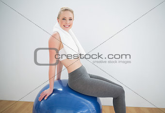 Smiling sporty blonde with towel around her neck sitting on exercise ball