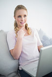 Casual thoughtful blonde sitting on couch using laptop