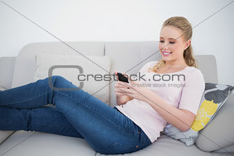 Casual smiling blonde using smartphone and lying on couch