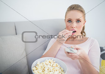 Casual astonished blonde holding remote and eating popcorn