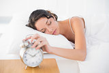 Sleepy casual brunette in white pajamas turning off her alarm clock