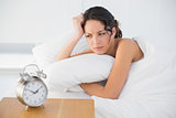 Frowning casual brunette in white pajamas looking at her alarm clock