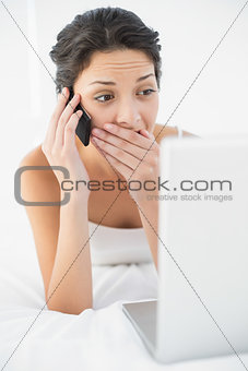 Surprised casual brunette in white pajamas making a phone call