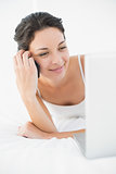Pleased casual brunette in white pajamas making a phone call and looking at her laptop