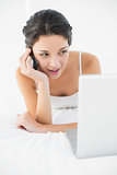 Cheerful casual brunette in white pajamas making a phone call and looking at her laptop