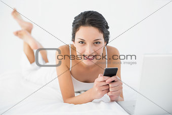 Delighted casual brunette in white pajamas using her mobile phone lying on her bed