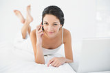 Pretty casual brunette in white pajamas making a phone call and smiling at camera
