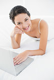 Smiling casual brunette in white pajamas using a laptop