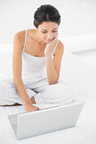 Concentrated casual brunette in white pajamas sitting on her bed and using a laptop