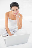 Cheerful casual brunette in white pajamas sitting on her bed and using a laptop