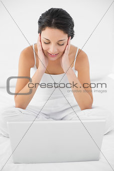 Dreamy casual brunette in white pajamas sitting on her bed and looking at her laptop