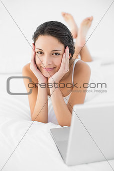 Happy casual brunette in white pajamas lying on her bed and looking at camera