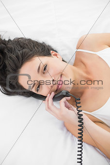 Pleased casual brunette in white pajamas calling with a telephone