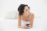 Thoughtful casual brunette in white pajamas holding a mobile phone