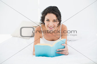 Smiling casual brunette in white pajamas relaxing on a bed reading a book