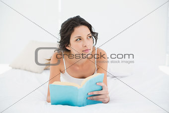 Thoughtful casual brunette in white pajamas relaxing on a bed reading a book