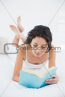 Calm casual brunette in white pajamas relaxing on a bed reading a book