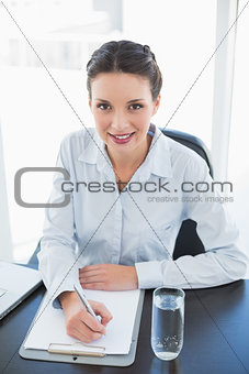 Cute stylish brunette businesswoman taking notes and looking at camera