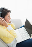 Pleased casual brunette in yellow cardigan listening to music while using a laptop