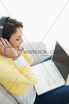 Pleased casual brunette in yellow cardigan listening to music while using a laptop