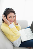 Cheerful casual brunette in yellow cardigan listening to music while using a laptop