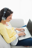 Calm casual brunette in yellow cardigan listening to music while using a laptop
