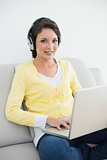 Smiling casual brunette in yellow cardigan listening to music while using a laptop