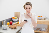 Thinking beautiful woman using her smartphone standing in the kitchen