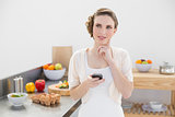Thoughtful pretty woman holding her smartphone standing in her kitchen