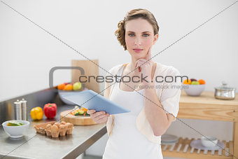 Thinking gorgeous woman standing in kitchen while holding her tablet