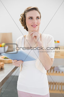 Smiling thinking woman holding her tablet standing in kitchen