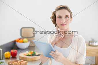 Young thoughtful woman standing in kitchen holing her tablet