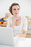 Thoughtful cute woman sitting in kitchen using her laptop