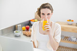 Cute woman drinking a glass of orange juice sitting in her kitchen in front of her notebook