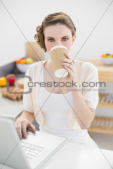 Beautiful woman drinking of disposable cup using her notebook in kitchen